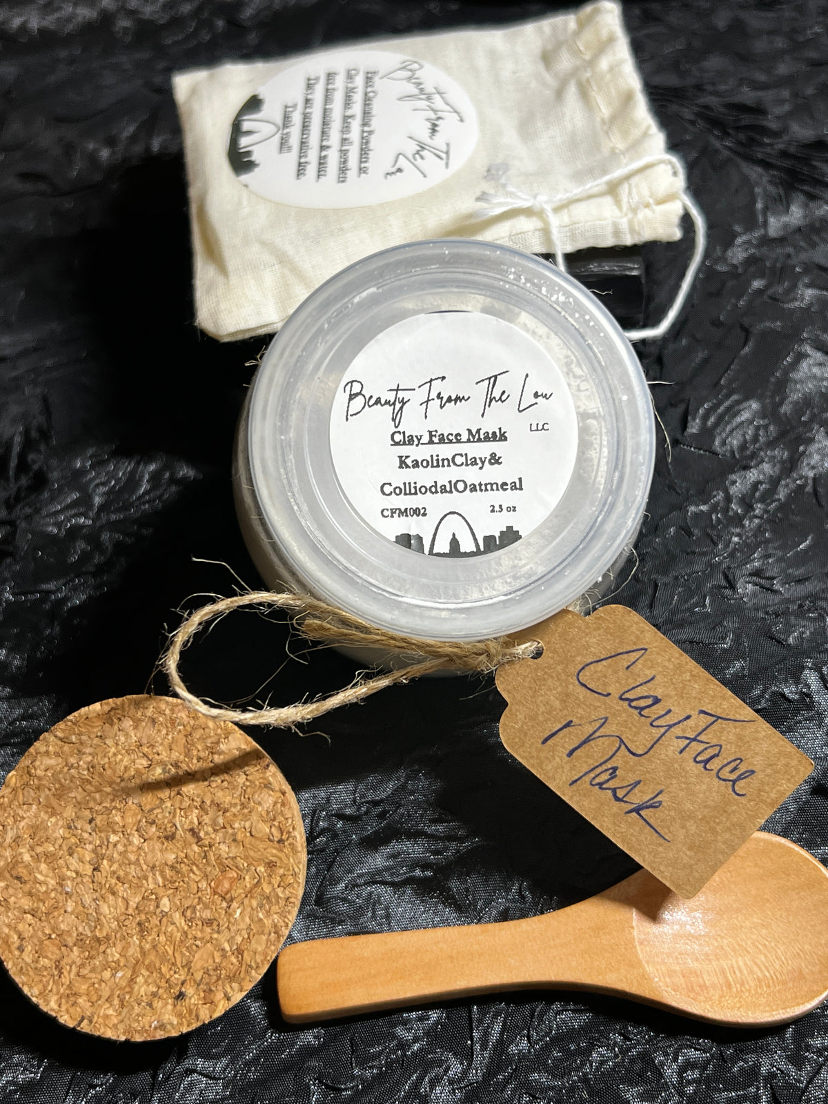 A jar of Clay Face Mask from Beauty From The Lou is displayed on a dark textured background with its accessories: a beige drawstring pouch, a wooden spoon, a cork and a paper tag.