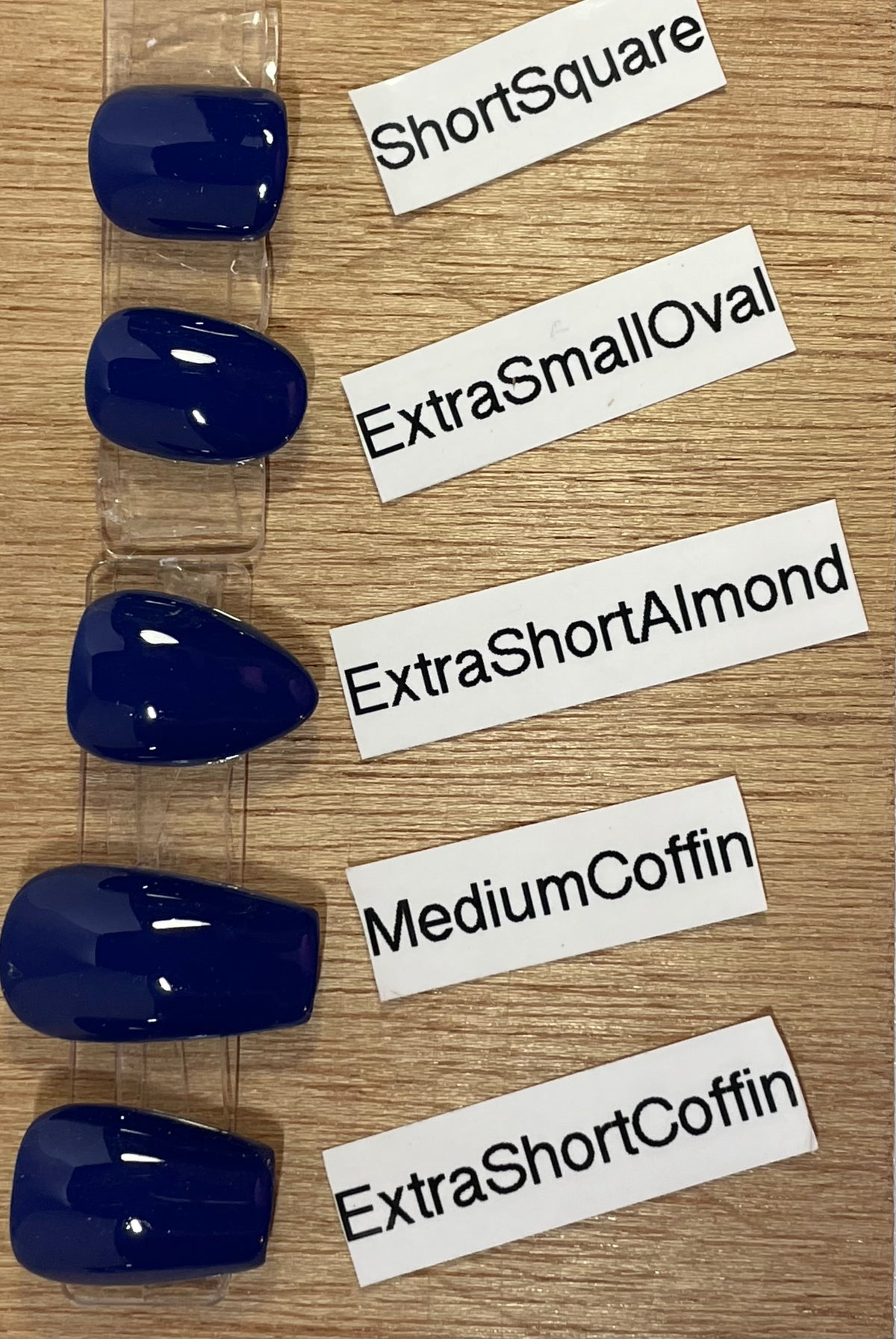 Five glossy dark blue coffin-shaped press-on nails displayed on a clear plastic strip, arranged vertically on a light wood surface with white labels reading ShortSquare, ExtraSmallOval, ExtraShortAlmond, MediumCoffin, and ExtraShortCoffin.