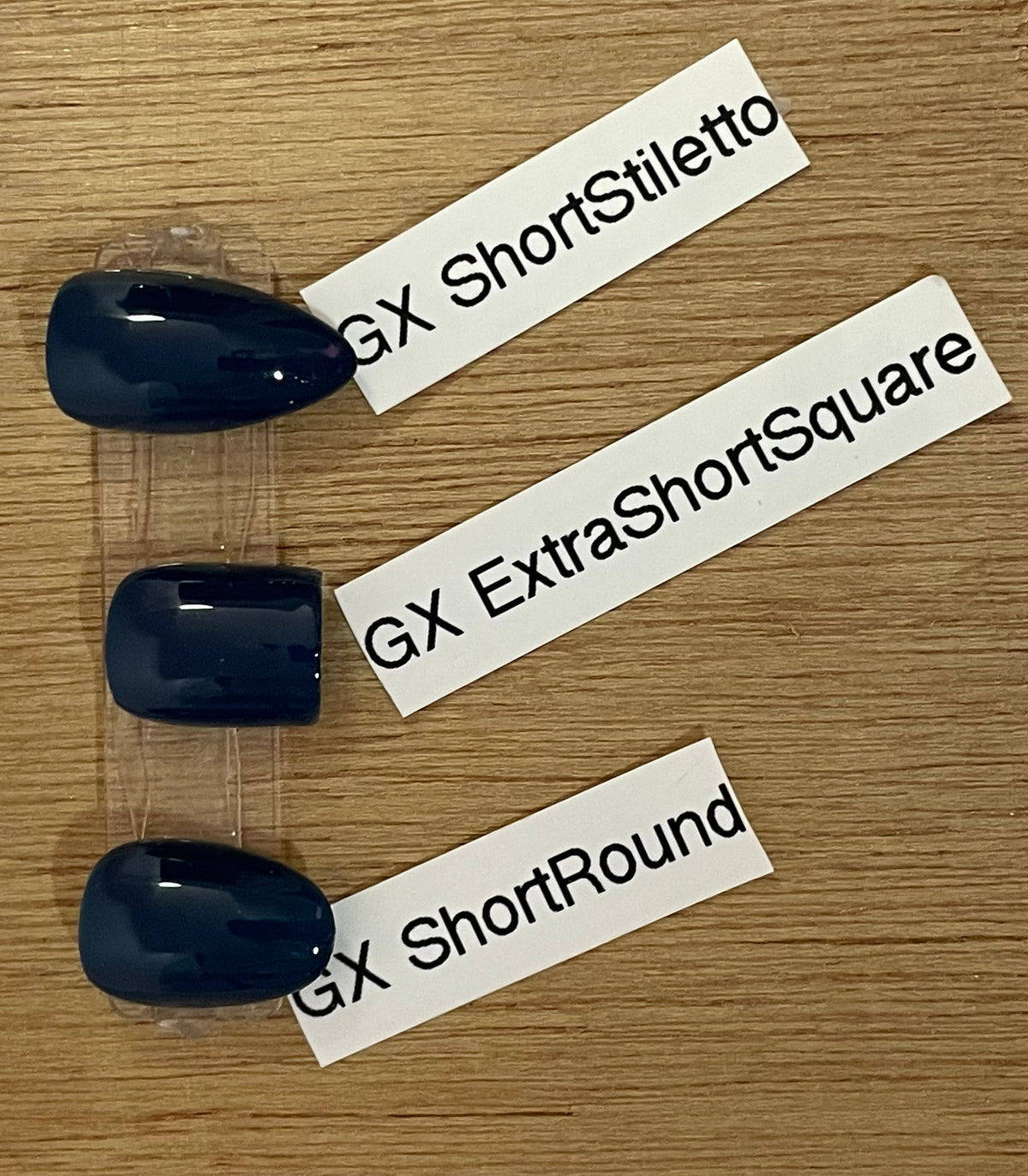 Three glossy dark blue press-on nails in different shapes (short stiletto, extra short square, short round) arranged vertically on a wooden surface with white labels pointing to each style.