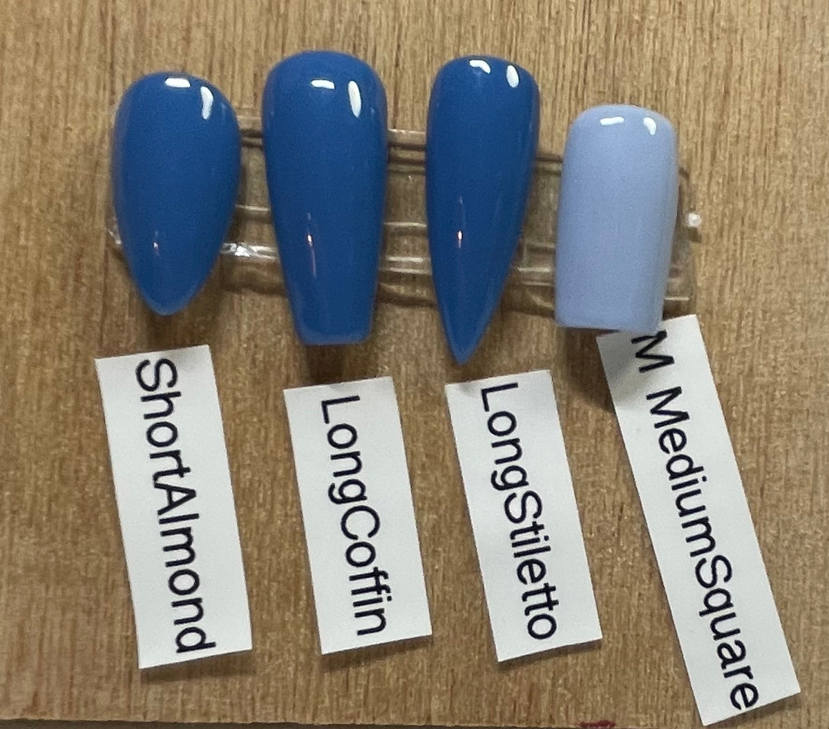 Four false nail tips in shades of blue and pale blue, labeled ShortAlmond, LongCoffin, LongStiletto, and M MediumSquare, displayed on a wooden surface.