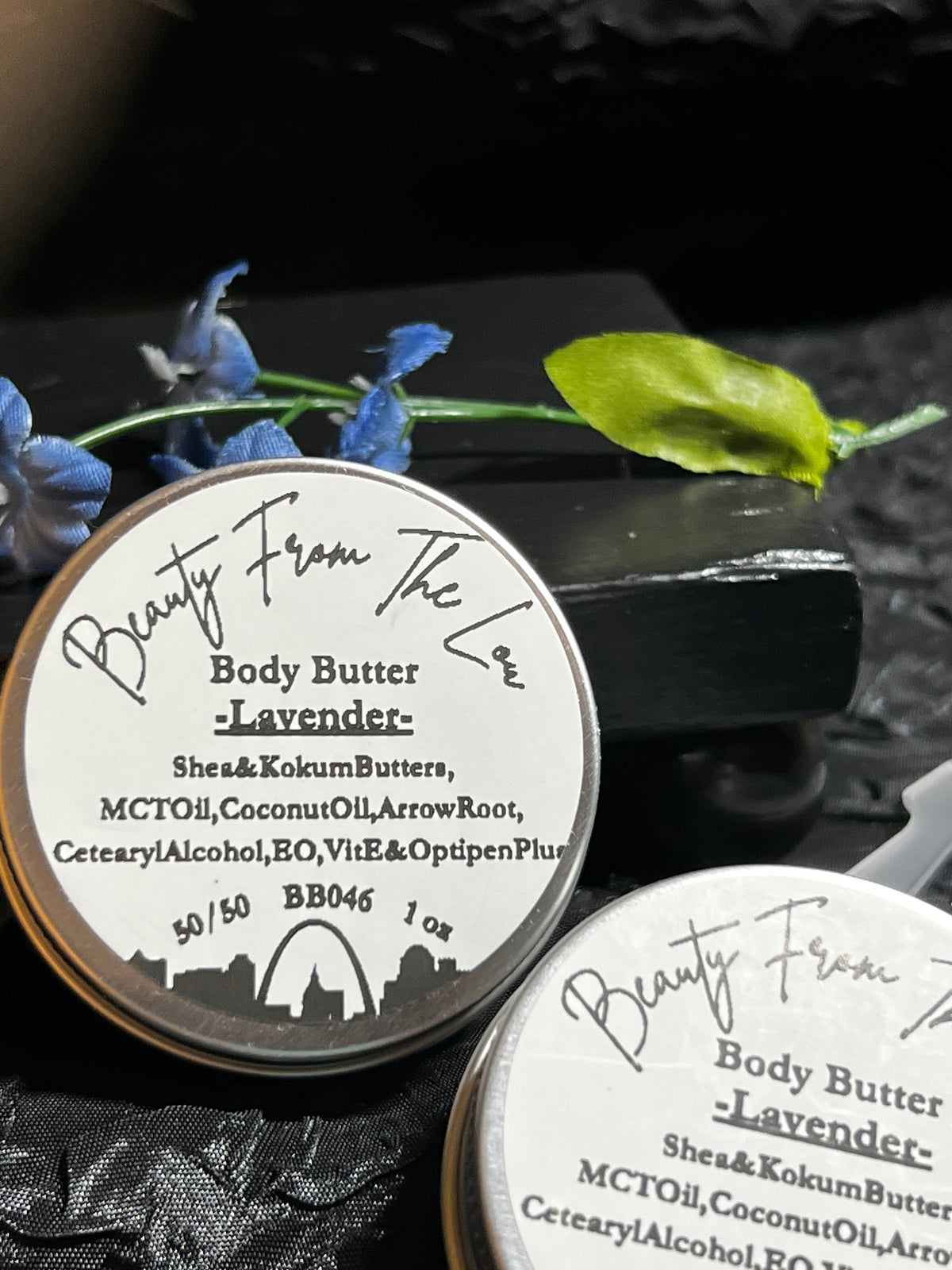 Two round metal tins of body butter labeled 'Beauty From The Lou' and 'Lavender' arranged on a dark textured surface with blue flowers and a green leaf as props.
