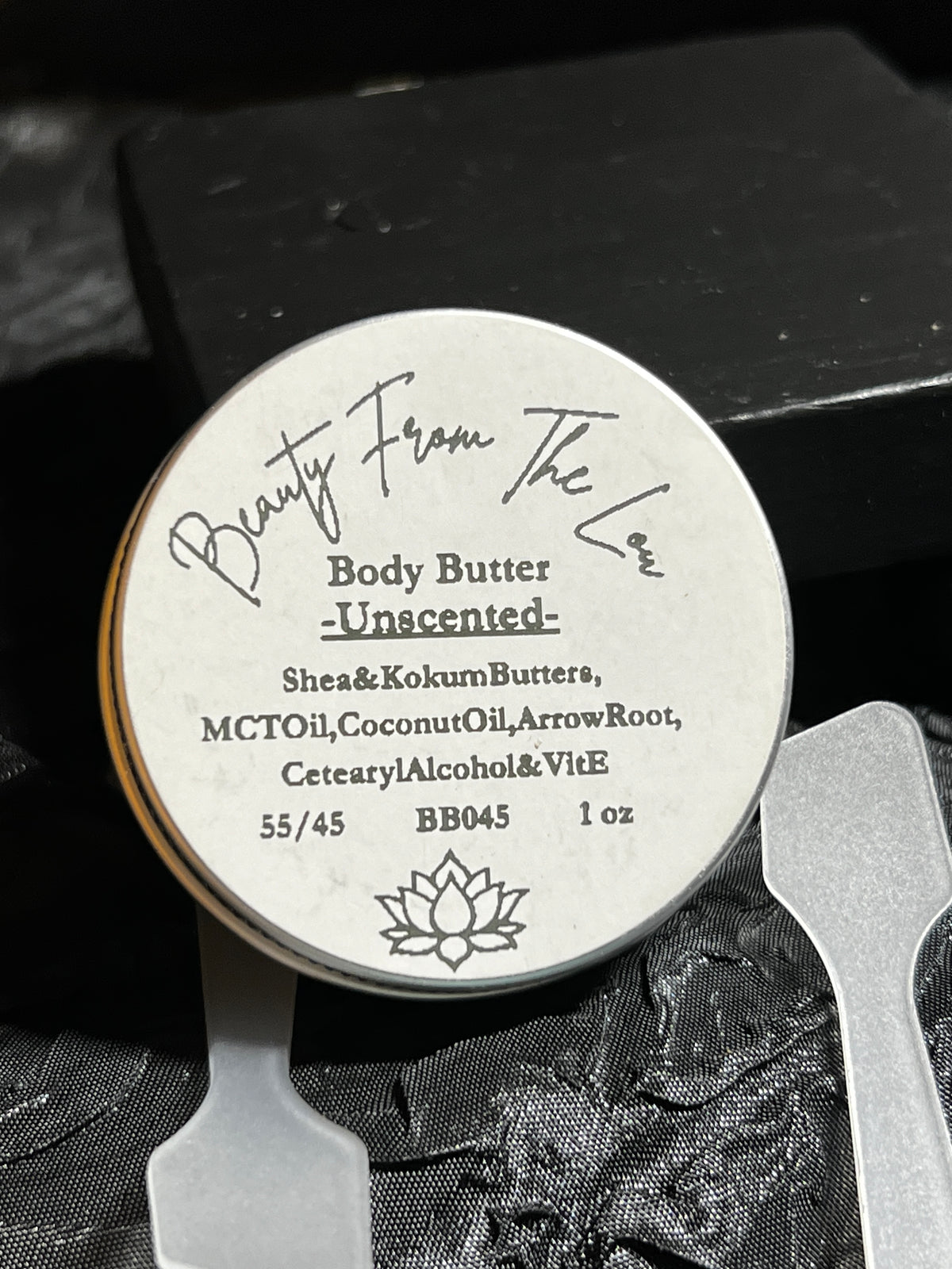 A round tin of Beauty From The Lou Body Butter, Unscented, with a white label and silver rim, displayed on a black textured surface with two white spatulas.
