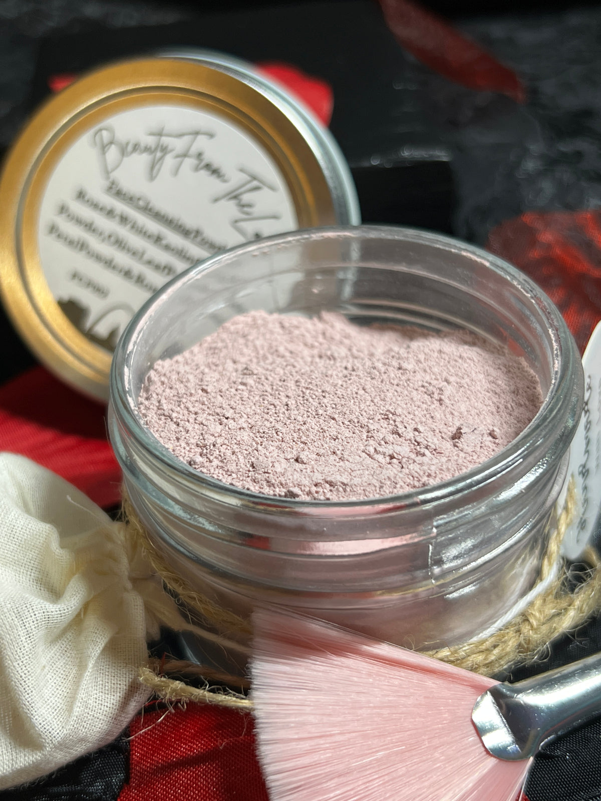 A small clear glass jar filled with light pink powder, shown with its silver lid in the background and a pink makeup brush in the foreground on a dark, textured surface.