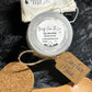 A jar of Clay Face Mask from Beauty From The Lou is displayed on a dark textured background with its accessories: a beige drawstring pouch, a wooden spoon, a cork and a paper tag.