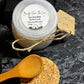 2.5 oz jar of a Face Mask made of Kaolin Clay & Olive Leaf Powder with a plastic lid and an extra lid made of cork and a wooden spoon.