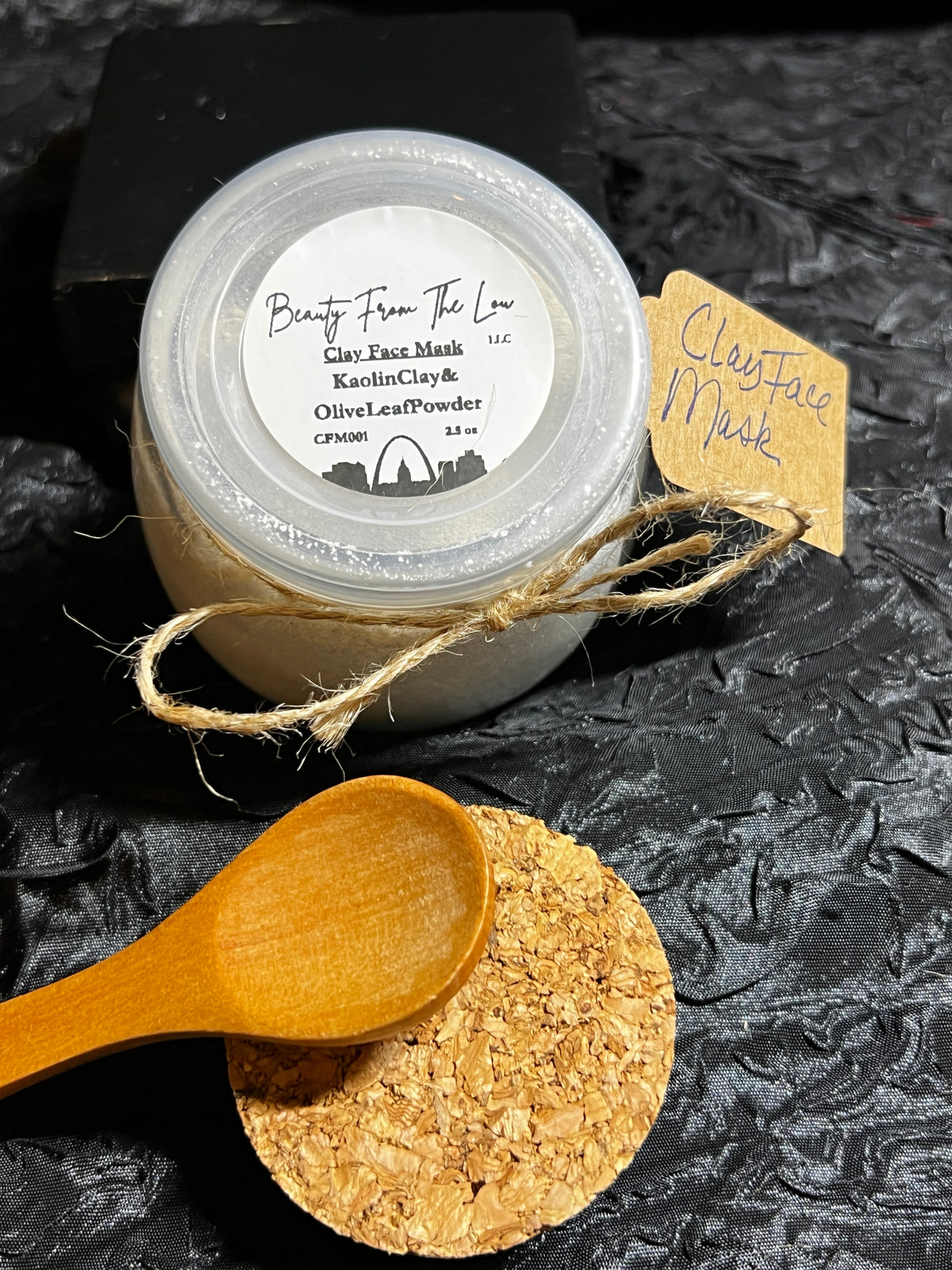 2.5 oz jar of a Face Mask made of Kaolin Clay & Olive Leaf Powder with a plastic lid and an extra lid made of cork and a wooden spoon.