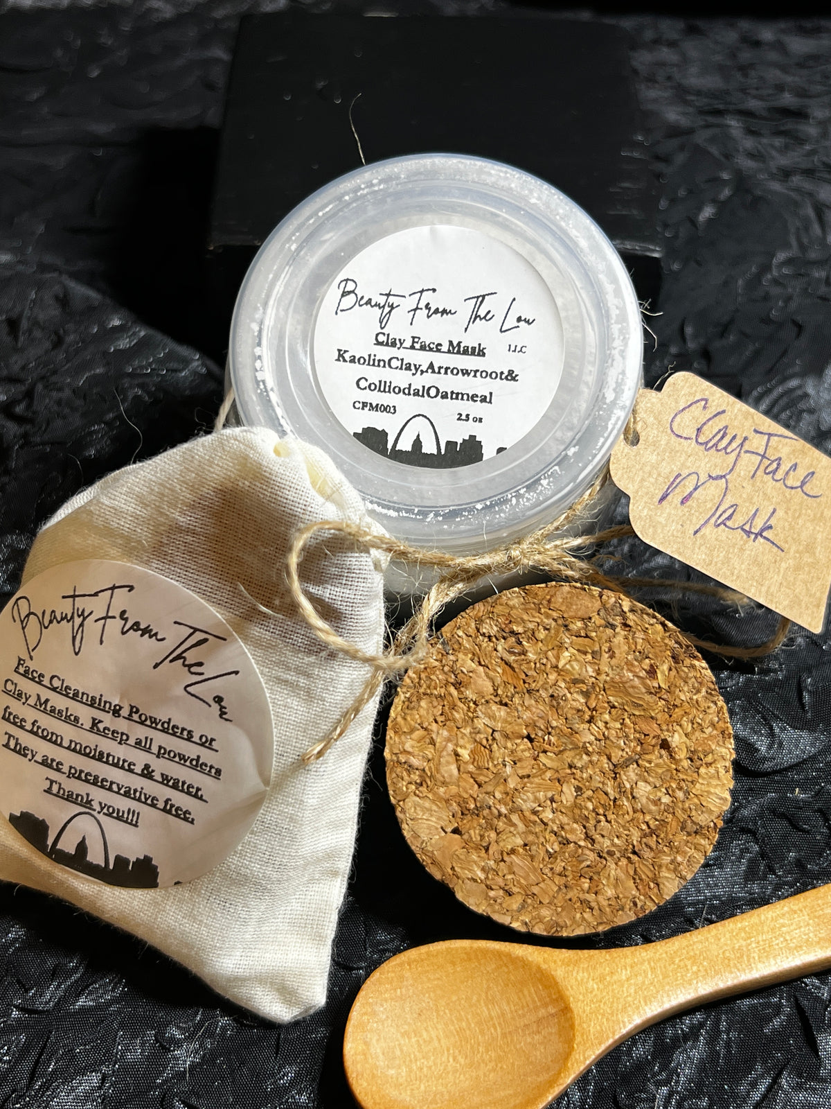 Beauty From The Lou clay face mask set featuring a small clear jar of clay face mask, a beige fabric drawstring pouch with a product label, a round cork stopper, and a wooden spoon on a dark textured background.
