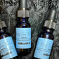 Three small 1/2 ounce dark glass dropper bottles of beard oil with light blue labels arranged on a textured black fabric background. The labels read 'Beauty From The Lou' and 'Beard Oil'.