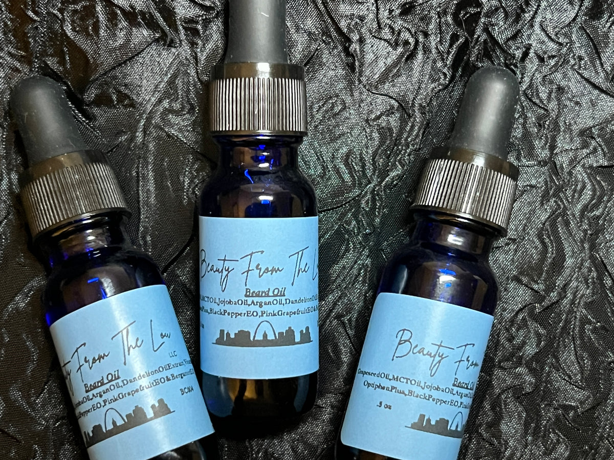 Three small 1/2 ounce dark glass dropper bottles of beard oil with light blue labels arranged on a textured black fabric background. The labels read 'Beauty From The Lou' and 'Beard Oil'.