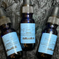 Three dark blue glass dropper bottles of beard oil with light blue labels, arranged on a dark textured fabric background. The labels read 'Beauty From The Lou' and 'Beard Oil'.