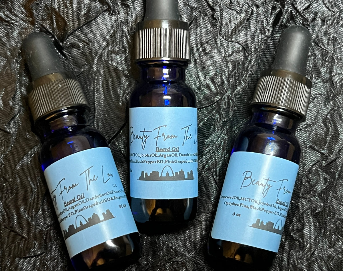 Three dark blue glass dropper bottles of beard oil with light blue labels, arranged on a dark textured fabric background. The labels read 'Beauty From The Lou' and 'Beard Oil'.